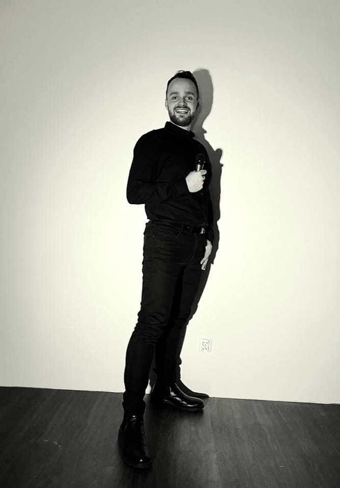 Uśmiechnięty mężczyzna w czarnym stroju, trzymający mikrofon, stojący na tle białej ściany z widocznym gniazdkiem elektrycznym i cieniem.