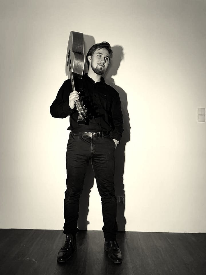 Czarno-białe zdjęcie muzyka z gitarą na ramieniu, ubrany na czarno, stojący na tle jasnej ściany i ciemnej podłogi.