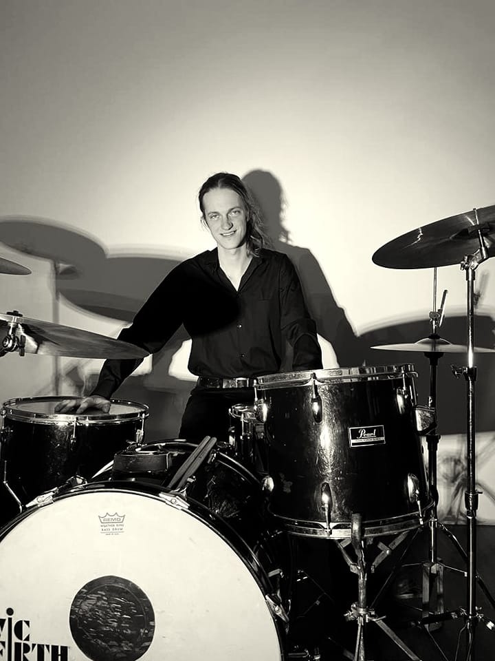 Portret perkusisty w czarnej koszuli siedzącego za zestawem perkusyjnym Pearl w monochromatycznej tonacji, z widocznym uśmiechem i długimi włosami.