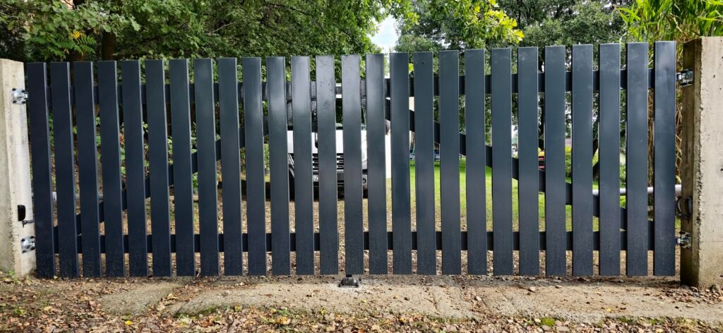 Nowoczesna brama wjazdowa z metalowymi sztachetami w kolorze grafitowym, zamontowana między betonowymi słupkami. Widoczny mechanizm otwierania bramy.