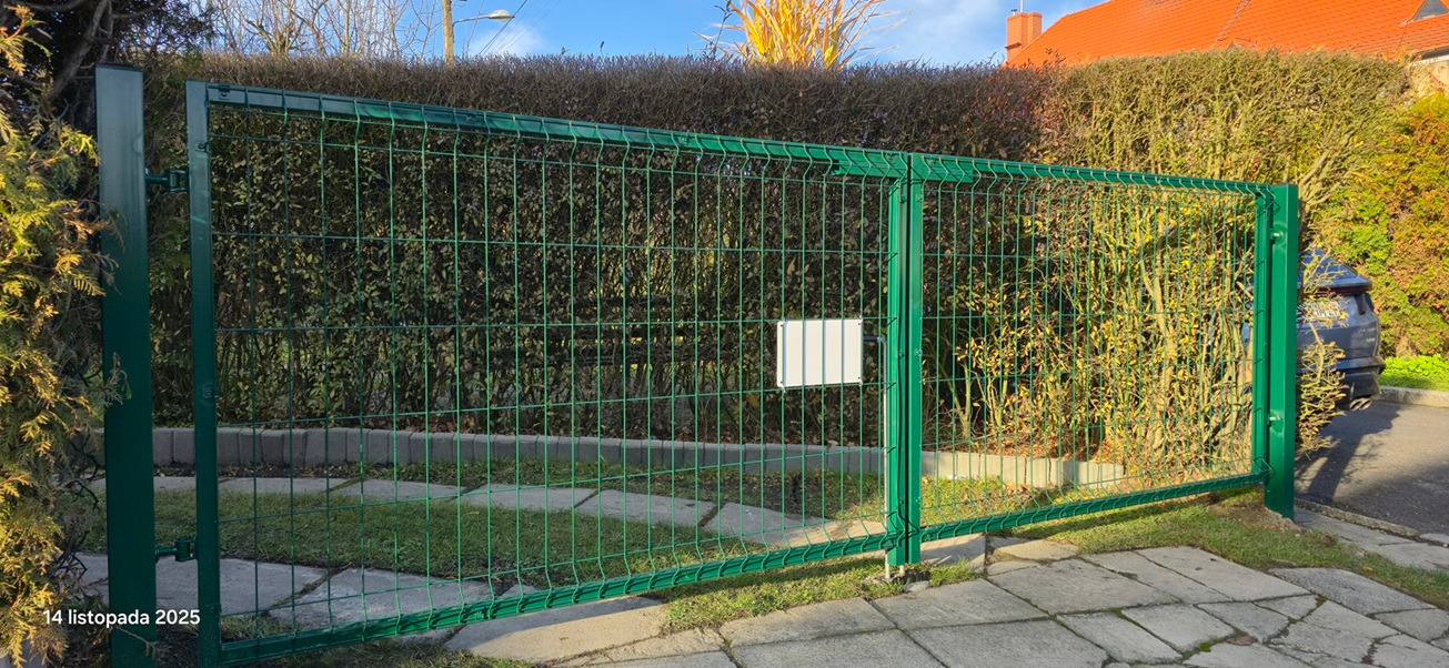Zielona brama wjazdowa z siatki panelowej na tle żywopłotu i kostki brukowej. Widoczny fragment samochodu za bramą. Brama dwuskrzydłowa z pustą białą tabliczką.