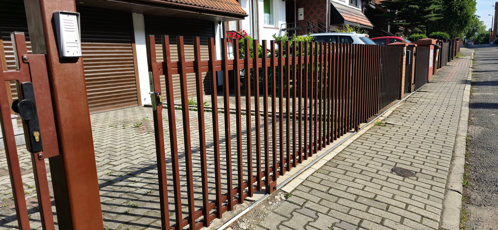Brązowa brama przesuwna z pionowych profili, widok z boku na tle kostki brukowej i zabudowań. Brama wyposażona w domofon i zamek.