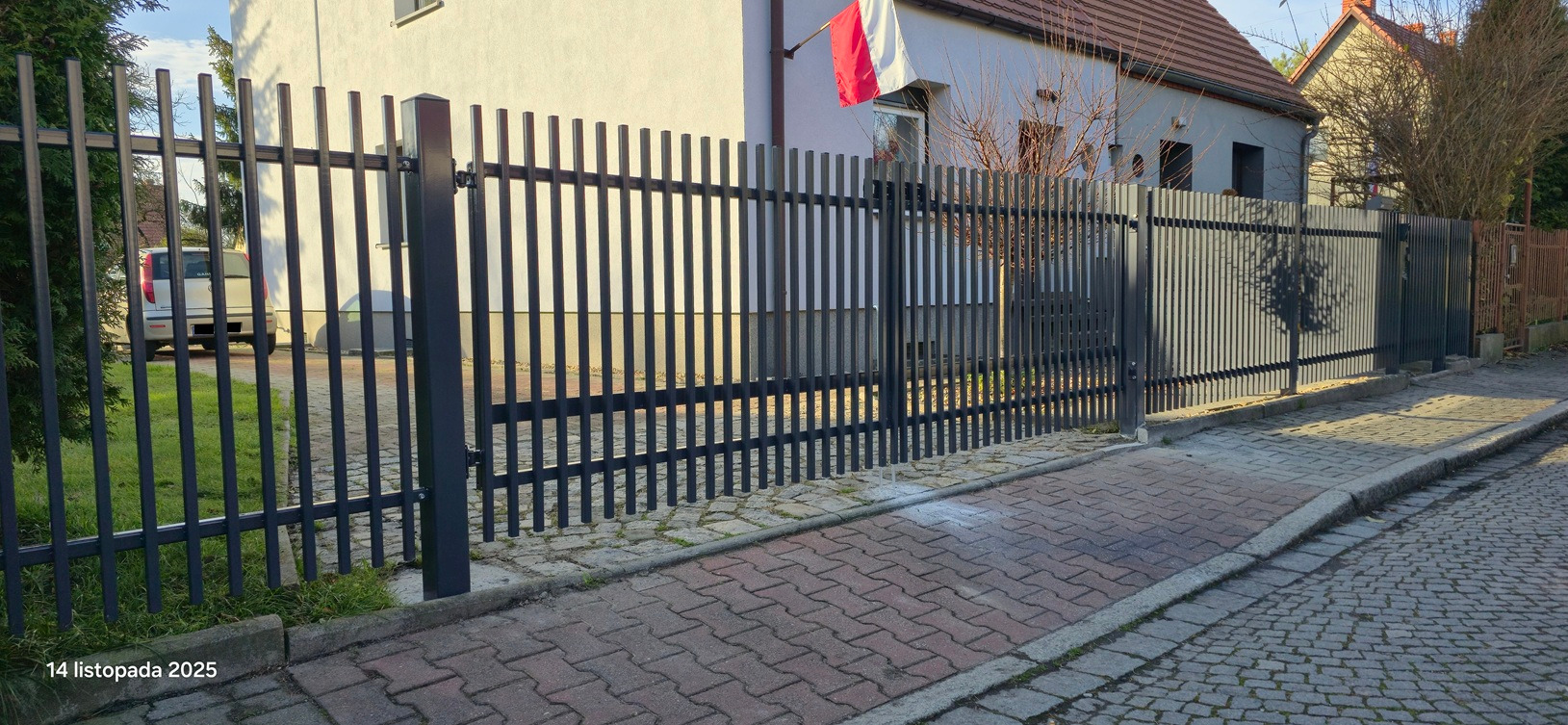 Nowoczesne, metalowe ogrodzenie panelowe w kolorze grafitowym, z bramą wjazdową. Widok z zewnątrz posesji, na tle domu z polską flagą. Kostka brukowa na pierwszym planie.