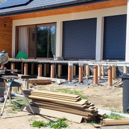 K-MONT - Budowa tarasu: widoczne fundamenty z rur, deski tarasowe ułożone na ziemi oraz piła stołowa na statywie przy nowoczesnym domu z panelami słonecznymi.