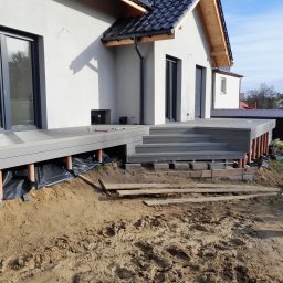 K-MONT - Montaż tarasu przy domu: widoczne podpory z rur PCV, czarna folia izolacyjna i fragment budynku z oknami tarasowymi.