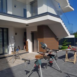 K-MONT - Montaż paneli elewacyjnych imitujących drewno na elewacji budynku, widoczna piła stołowa i drabina.