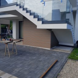 K-MONT - Montaż paneli elewacyjnych imitujących drewno pod schodami zewnętrznymi z balustradą szklaną; widoczne narzędzia i drabina na tle kostki brukowej.