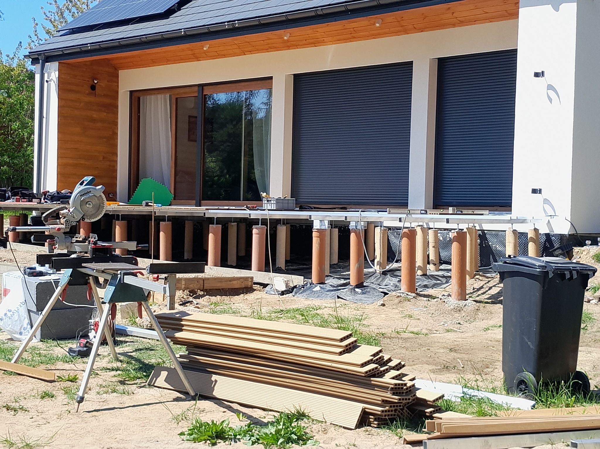 Budowa tarasu: widoczne fundamenty z rur, deski tarasowe ułożone na ziemi oraz piła stołowa na statywie przy nowoczesnym domu z panelami słonecznymi.