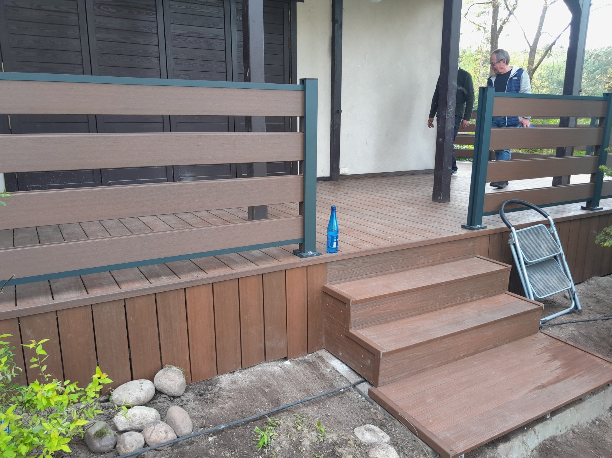 Nowoczesny taras z brązowych desek kompozytowych z niską balustradą, schodami i widoczną butelką wody mineralnej oraz osobami w tle.