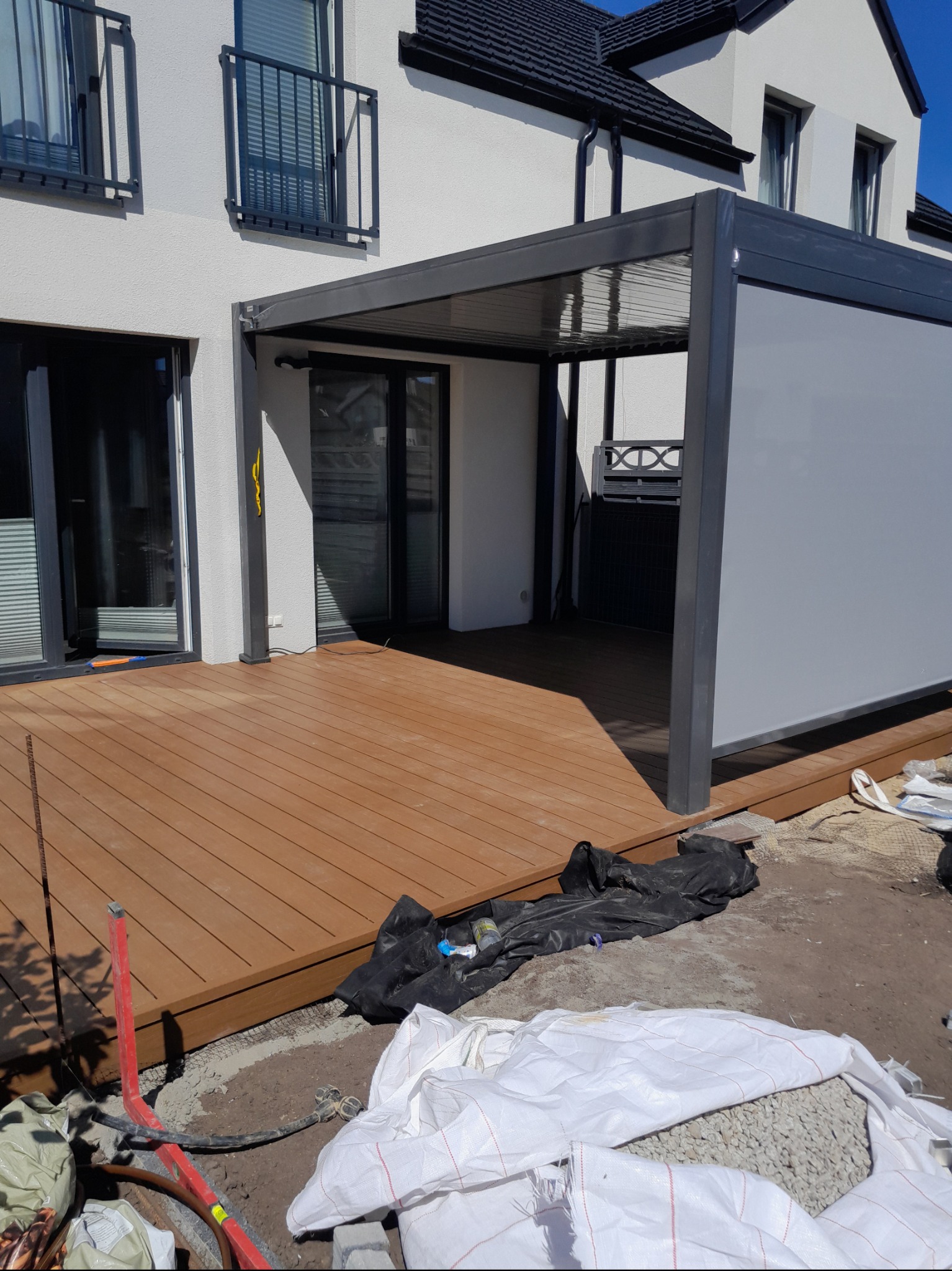 Montaż tarasu z desek kompozytowych przy nowoczesnym domu z pergolą, widoczne worki z kruszywem i narzędzia budowlane.