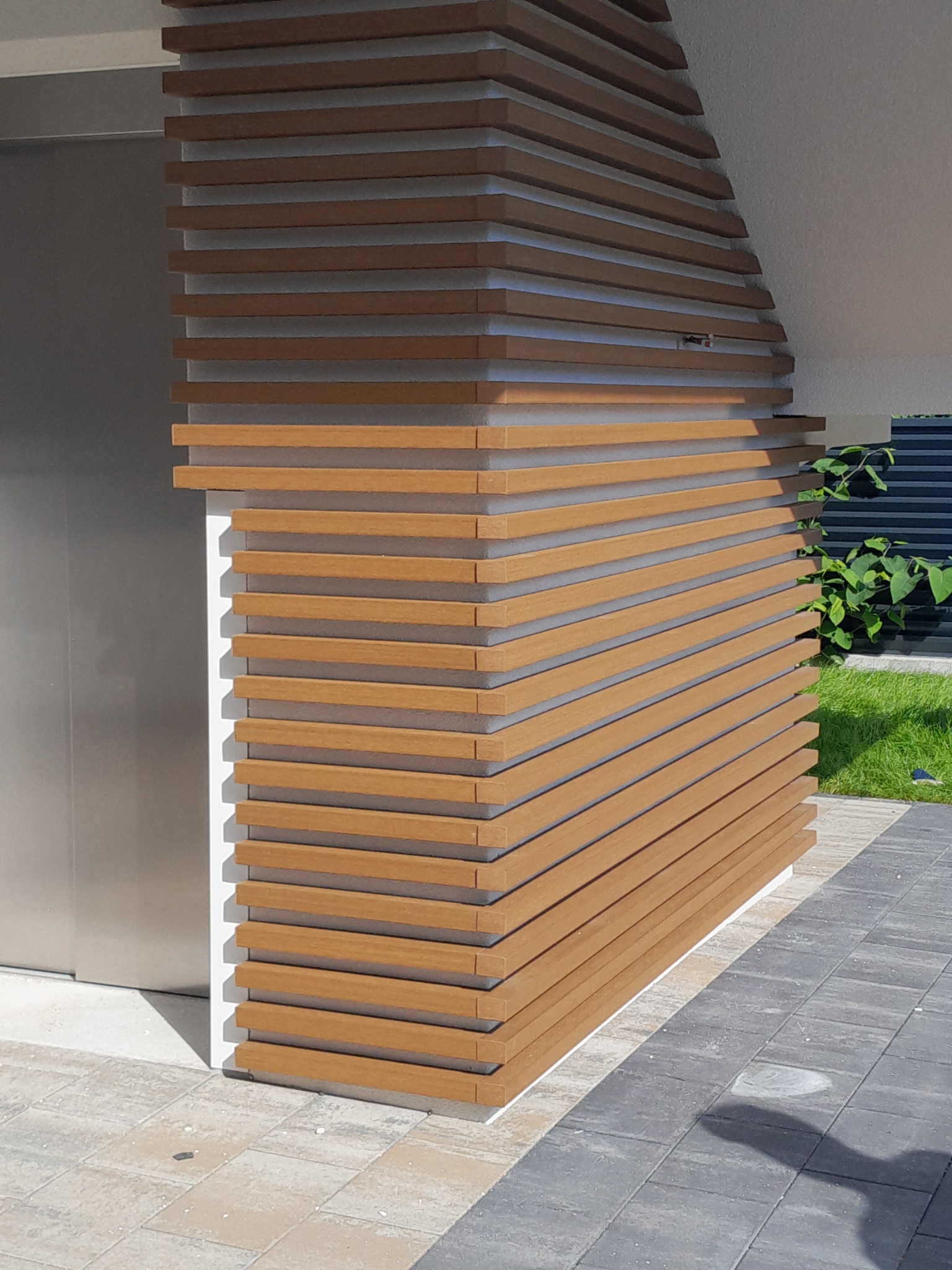 Nowoczesna elewacja z poziomych listew imitujących drewno, tworząca ażurową osłonę narożnika budynku, widoczna z poziomu kostki brukowej.