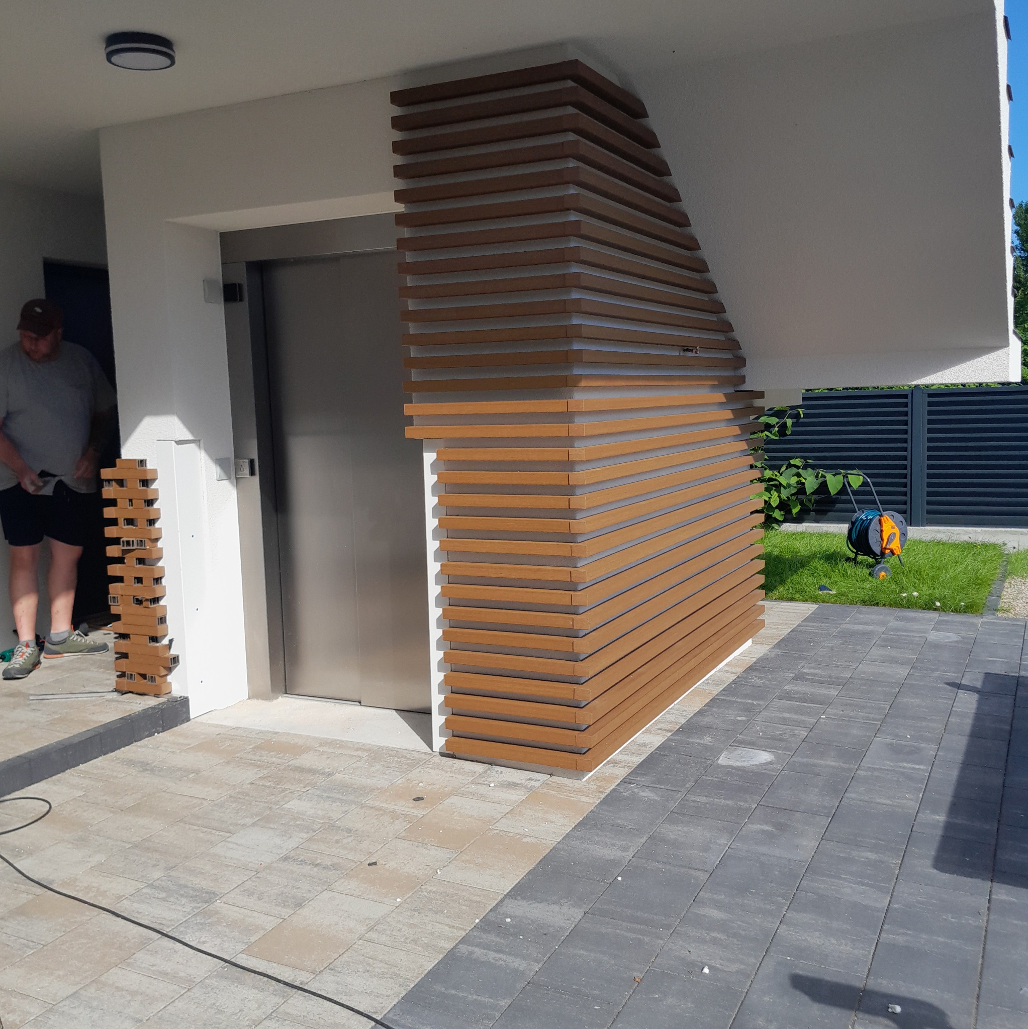 Montaż pionowej osłony z desek kompozytowych przy wejściu do budynku, widoczny pracownik z deskami i narzędziami.