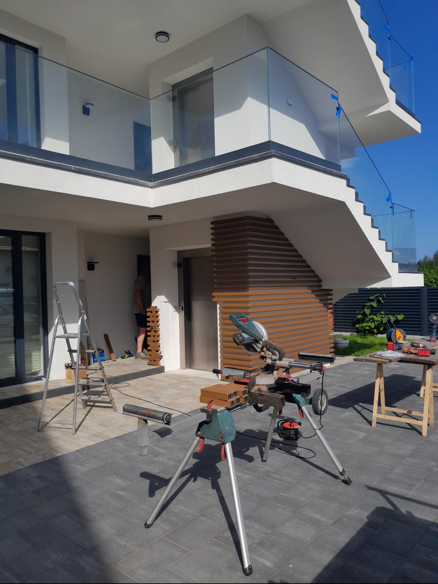 Montaż paneli elewacyjnych imitujących drewno na elewacji budynku, widoczna piła stołowa i drabina.