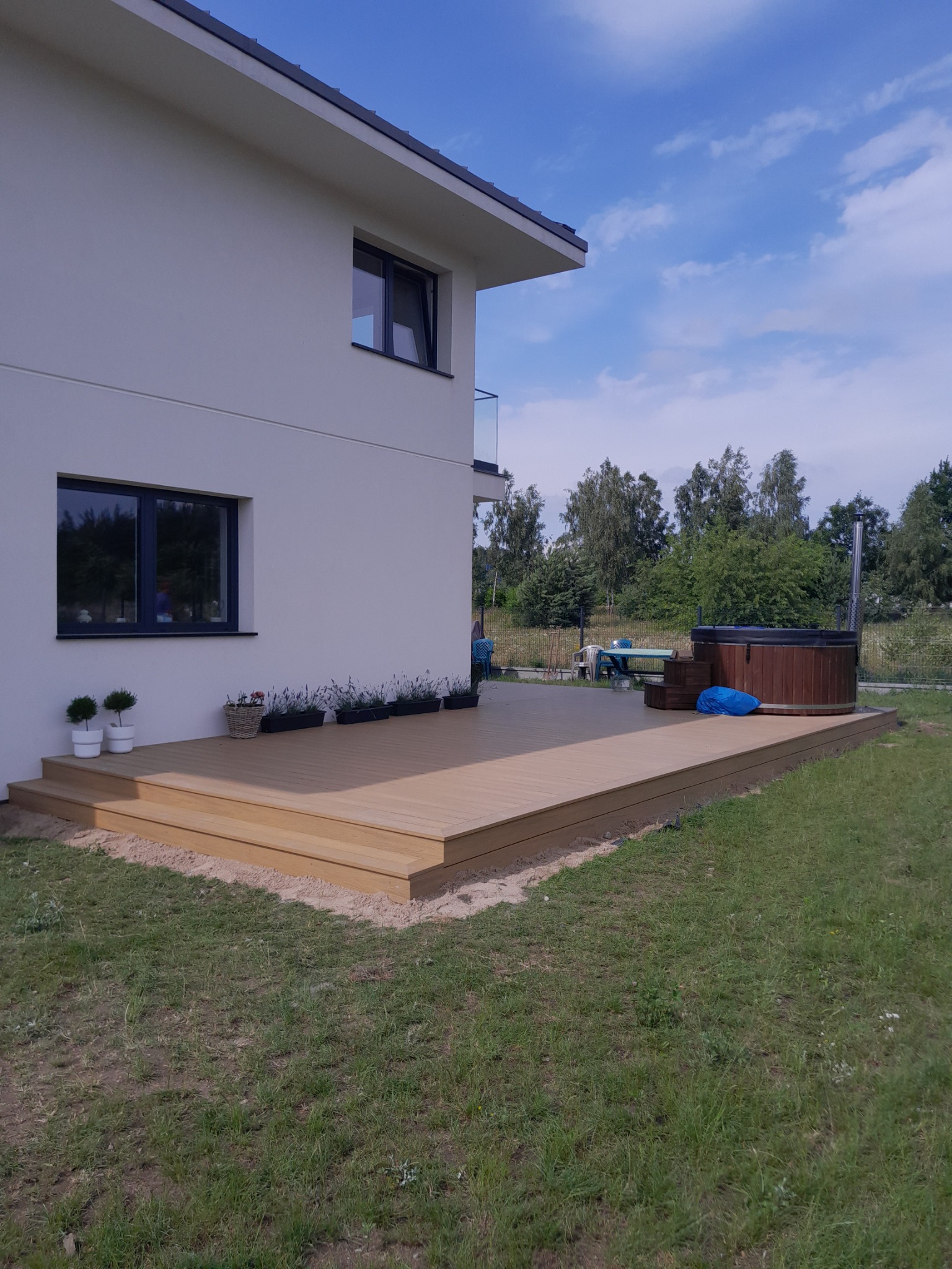 Nowoczesny taras przydomowy z drewnopodobnych desek, z widoczną balia ogrodową i roślinami w donicach przy ścianie budynku.