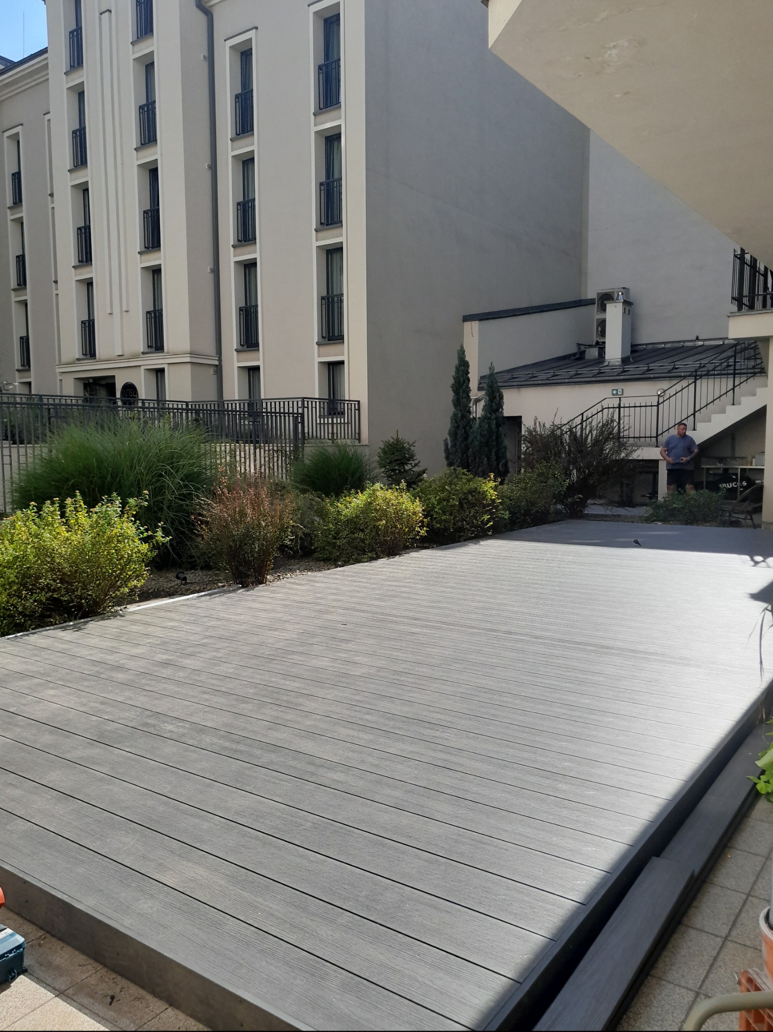 Szary taras kompozytowy z delikatnym usłojeniem, otoczony zielenią, z widocznym fragmentem budynku mieszkalnego i mężczyzną w tle.