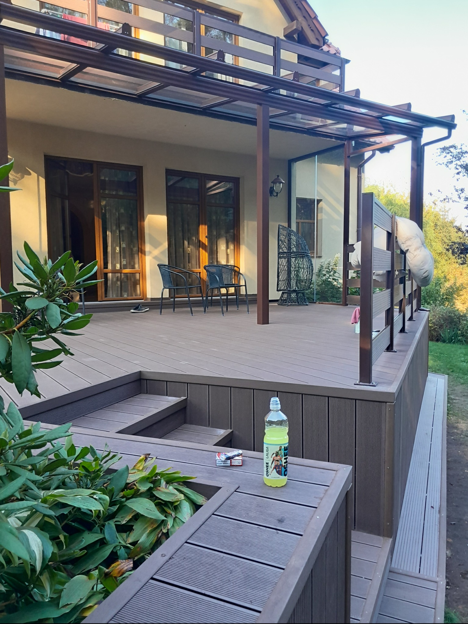 Zadaszony taras kompozytowy z brązowymi balustradami, schodkami i wbudowaną donicą z roślinami, butelką napoju i paczką papierosów na krawędzi.