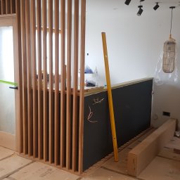 BUDEXJAR - Montaż drewnianej ścianki działowej z pionowych listew, widoczny poziomica, instalacja elektryczna w ścianie i lampa wisząca w folii ochronnej.