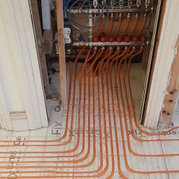 BUDEXJAR - Montaż ogrzewania podłogowego: pomarańczowe rury grzewcze ułożone na białej macie izolacyjnej z widocznym rozdzielaczem i przewodami elektrycznymi.