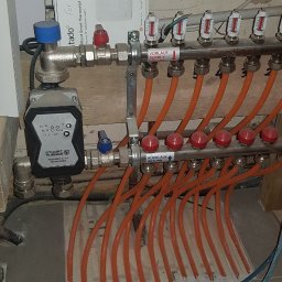 BUDEXJAR - Rozdzielacz ogrzewania podłogowego z przewodami w kolorze pomarańczowym, zaworami z czerwonymi pokrętłami, pompą obiegową Stuart Turner i pudełkiem termostatu Tado w tle, widoczne oznaczenia...