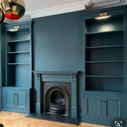 BUDEXJAR - Elegancki salon z zabudowanymi regałami i kominkiem w odcieniach morskiej zieleni, parkiet w jodełkę, oświetlenie punktowe nad półkami, minimalistyczny design.