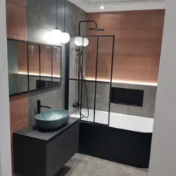 Nowoczesna łazienka z umywalką w kolorze morskim, kabiną prysznicową z czarną ramą i płytkami w odcieniach szarości i terakoty, oświetlona lampami wiszącymi.