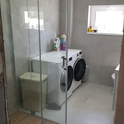 BUDEXJAR - Wykończona łazienka z kabiną prysznicową ze szkła, pralką i koszem na pranie. Ściany i podłoga wyłożone płytkami w odcieniach szarości. Widoczne okno z dekoracjami.