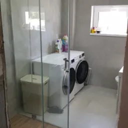 Wykończona łazienka z kabiną prysznicową ze szkła, pralką i koszem na pranie. Ściany i podłoga wyłożone płytkami w odcieniach szarości. Widoczne okno z dekoracjami.