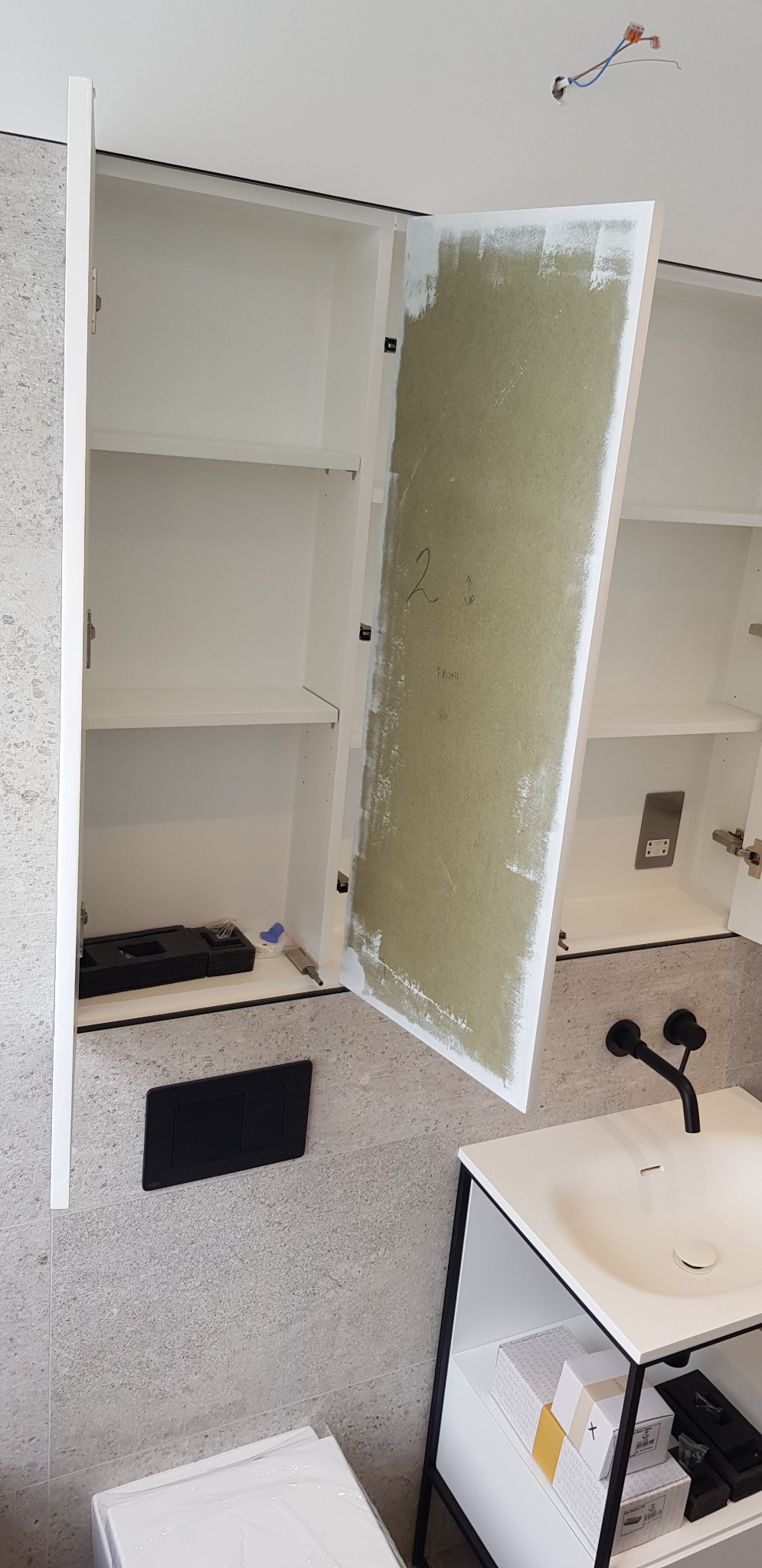 Otwarta szafka łazienkowa z niedomalowanymi drzwiczkami, widoczne akcesoria montażowe, czarna armatura i ceramika sanitarna na tle szarej ściany z płytek.