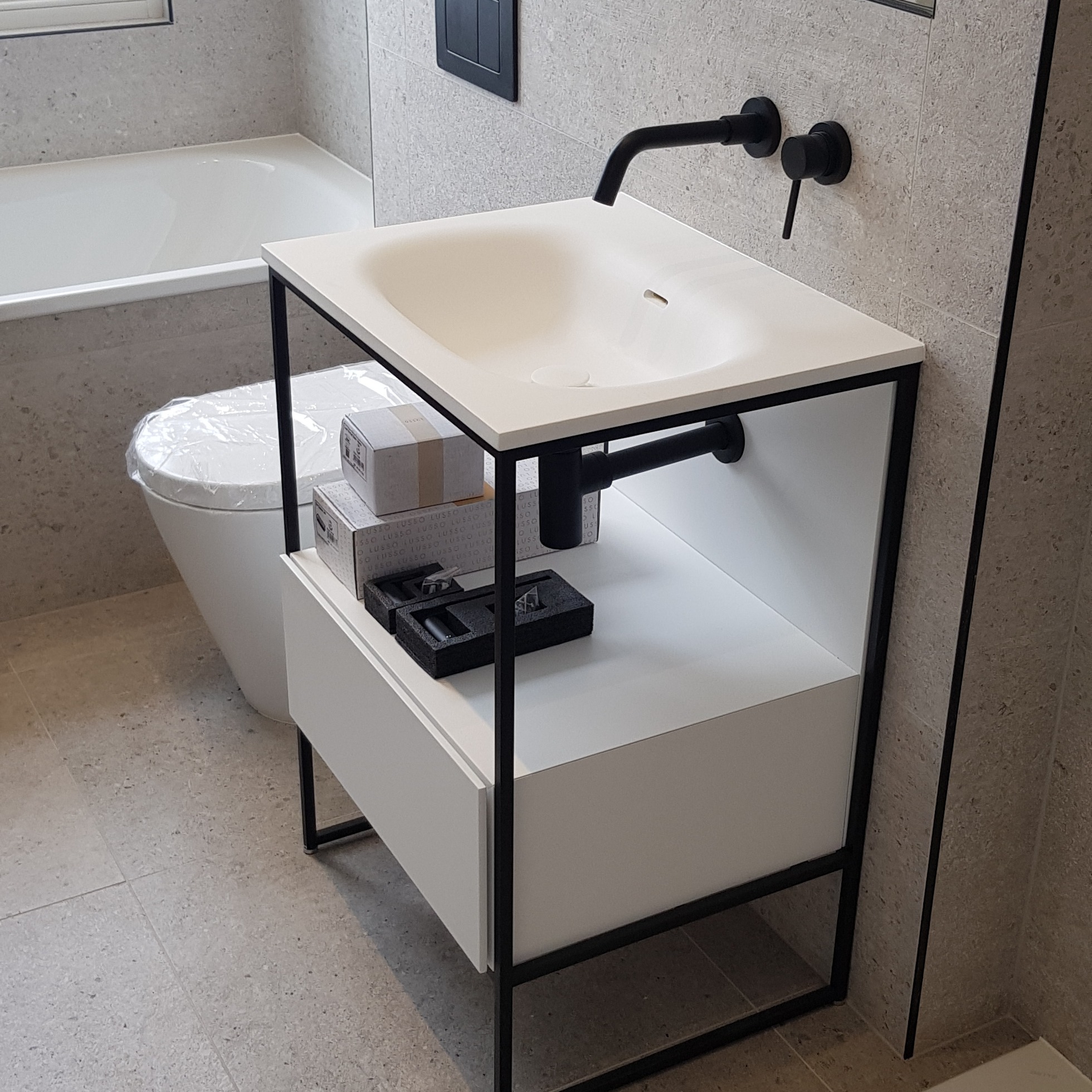 Nowoczesna łazienka z umywalką nablatową o białym blacie i czarnym stelażu, czarna bateria umywalkowa, toaleta w folii ochronnej i pudełka z akcesoriami na półce, szare płytki na ścianach i podłodze.