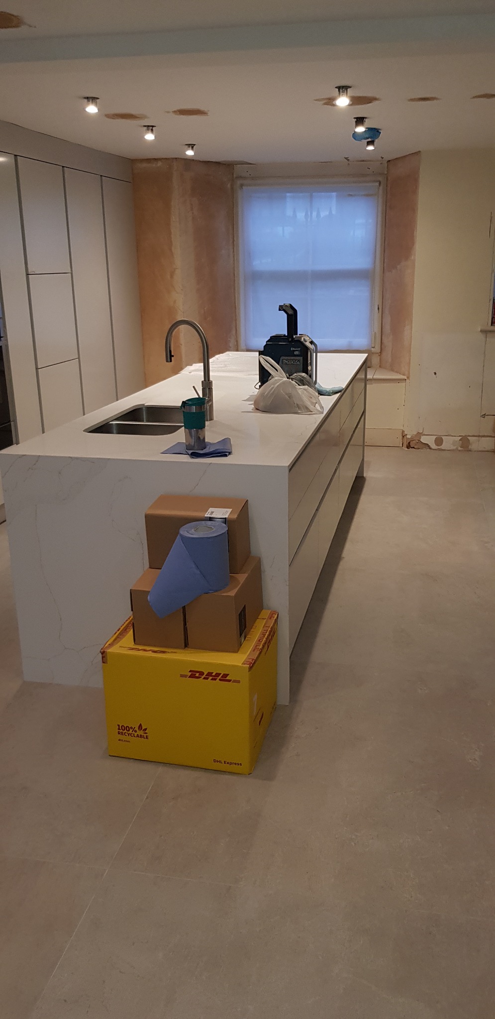 Niedokończona kuchnia z wyspą i stosem paczek DHL na podłodze, widoczne ślady po remoncie na ścianach i suficie.