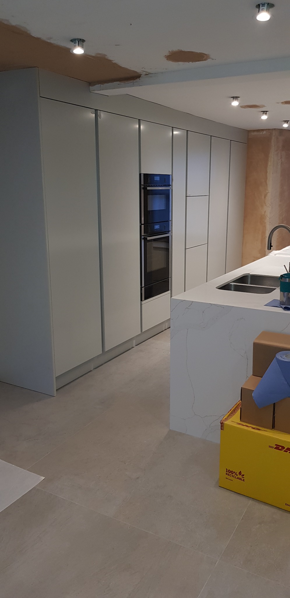 Nowoczesna kuchnia w trakcie wykończenia: jasne, minimalistyczne szafki, marmurowy blat wyspy, wbudowany piekarnik, surowy sufit z widocznymi śladami po instalacji oświetlenia, kartony na podłodze.