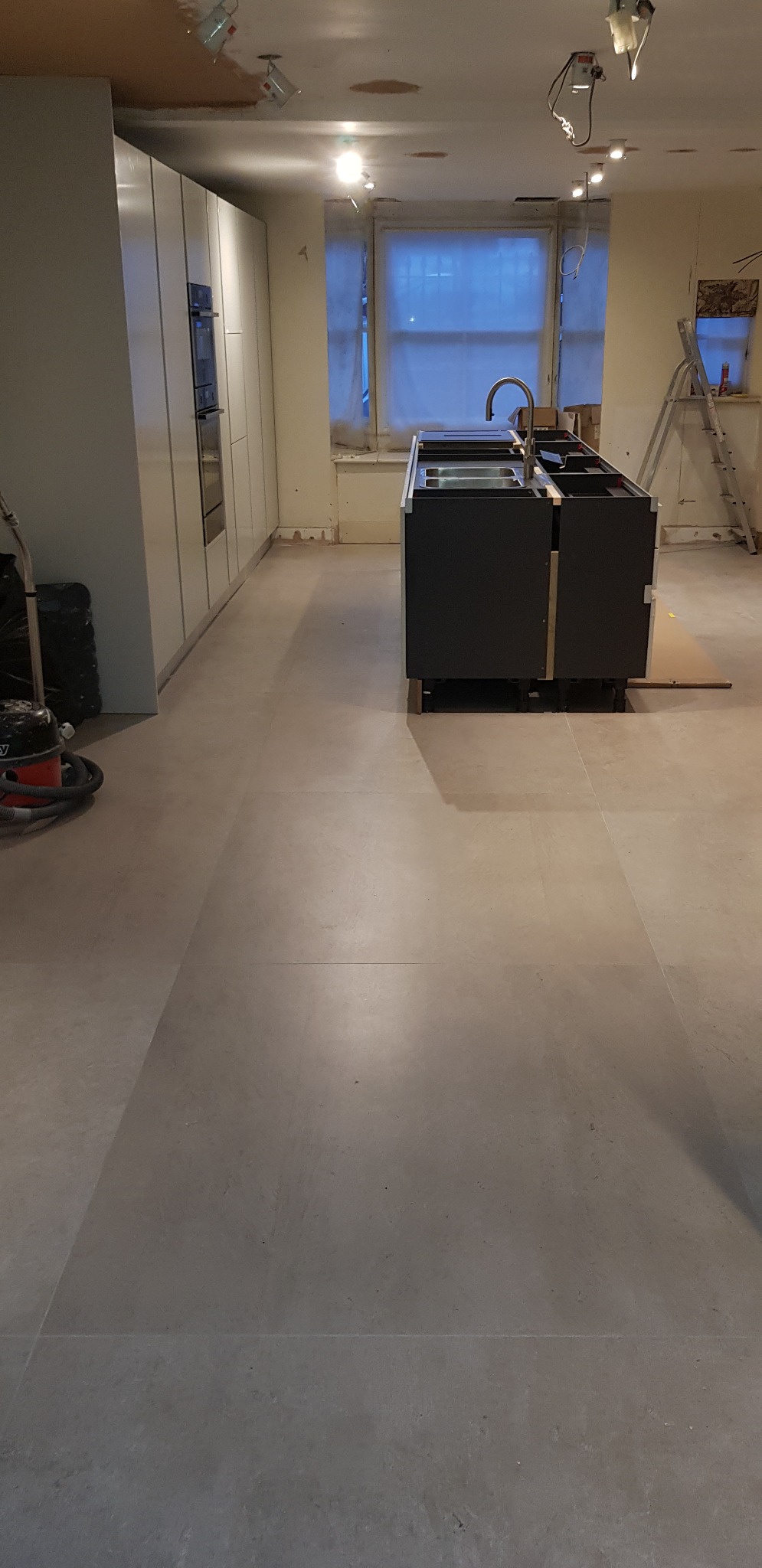 Montaż wyspy kuchennej z blatem i zlewem w nowym wnętrzu, widoczne białe szafki kuchenne, szara podłoga, drabina i odkurzacz przemysłowy w tle.