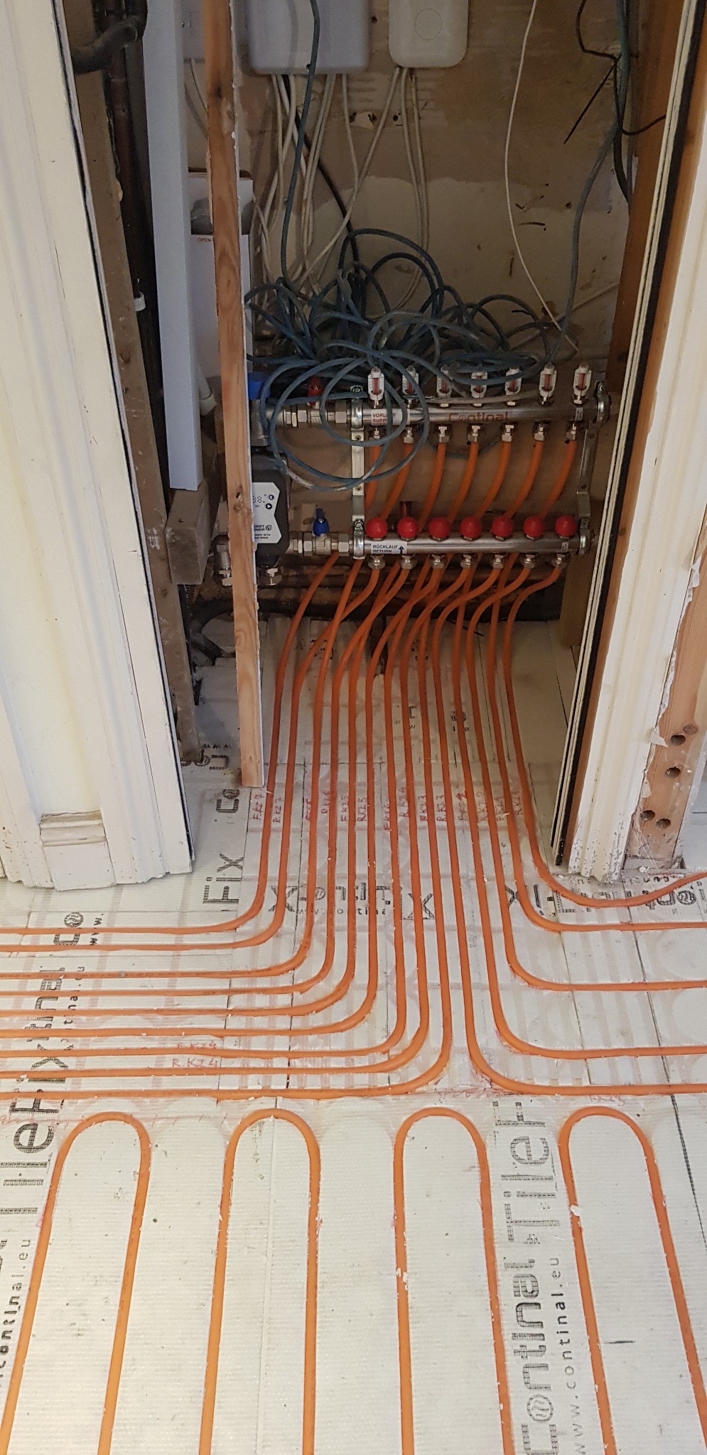 Montaż ogrzewania podłogowego: pomarańczowe rury grzewcze ułożone na białej macie izolacyjnej z widocznym rozdzielaczem i przewodami elektrycznymi.