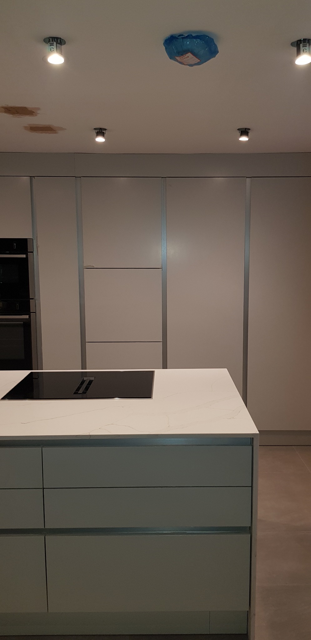 Nowoczesna kuchnia z szarymi szafkami, białą wyspą z czarną płytą indukcyjną i zabezpieczonym folią oświetleniem sufitowym.