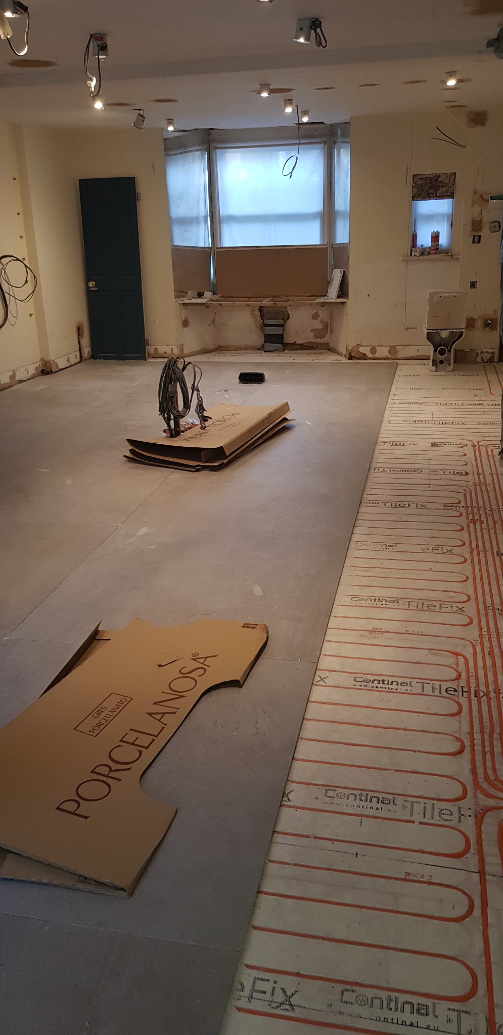 Przygotowanie do położenia płytek podłogowych w łazience, widoczne ogrzewanie podłogowe, kartony po płytkach Porcelanosa oraz narzędzia.