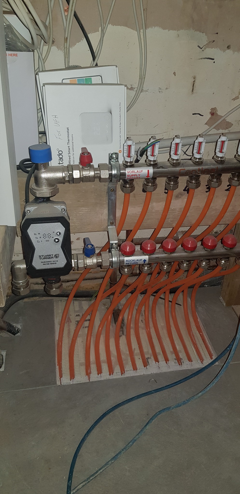 Rozdzielacz ogrzewania podłogowego z przewodami w kolorze pomarańczowym, zaworami z czerwonymi pokrętłami, pompą obiegową Stuart Turner i pudełkiem termostatu Tado w tle, widoczne oznaczenia...