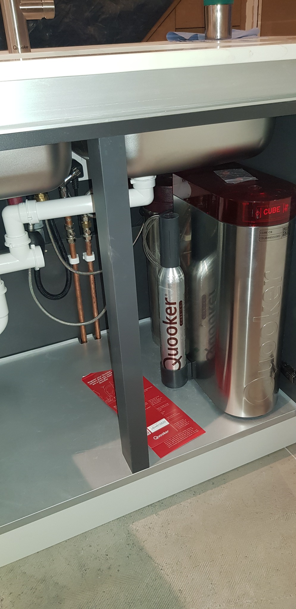 Montaż systemu Quooker Cube pod zlewem kuchennym, widoczne rury, zbiornik i butla CO2, czerwona ulotka informacyjna na dnie szafki.