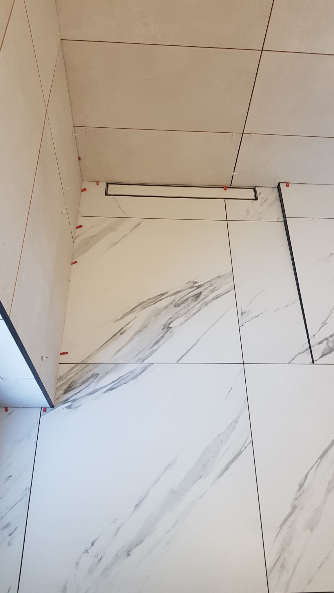 Biała łazienka z marmurkową podłogą w trakcie wykańczania. Widoczny odpływ liniowy i krzyżyki dystansowe między płytkami. Ułożone płytki na ścianach i podłodze.