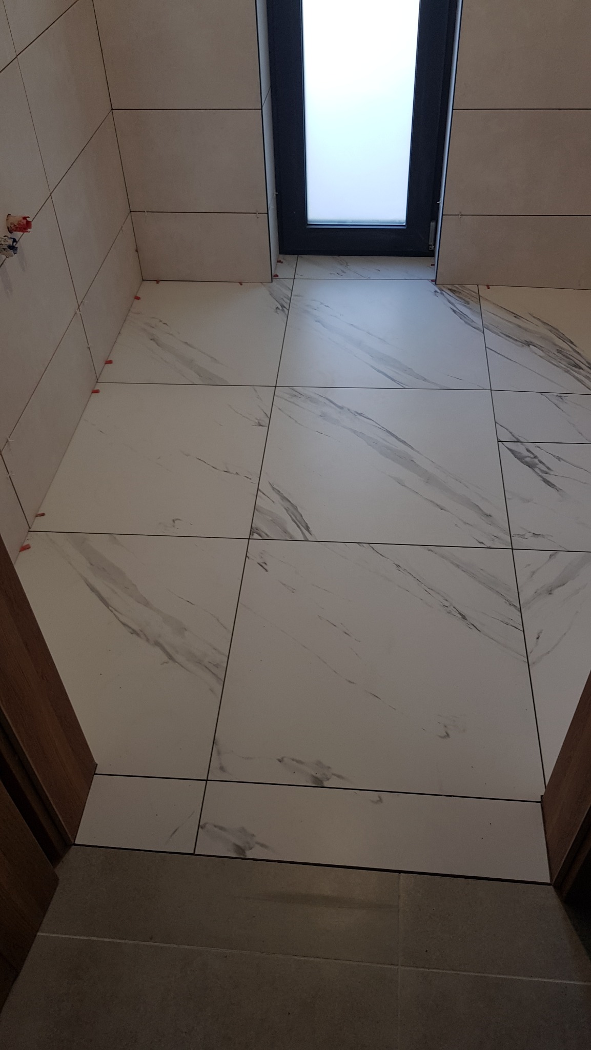 Układanie płytek podłogowych imitujących marmur, z widocznymi krzyżykami dystansowymi i oknem dachowym. Widok z góry na fragment łazienki w trakcie wykańczania.