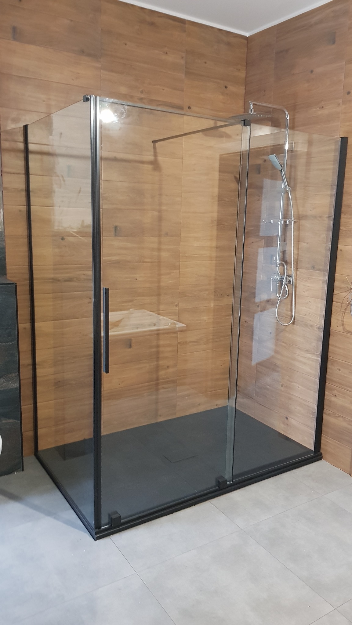 Nowoczesna kabina prysznicowa typu walk-in z czarnymi profilami, drewnopodobne płytki na ścianach, szara podłoga. Minimalistyczny design z półką wewnątrz.