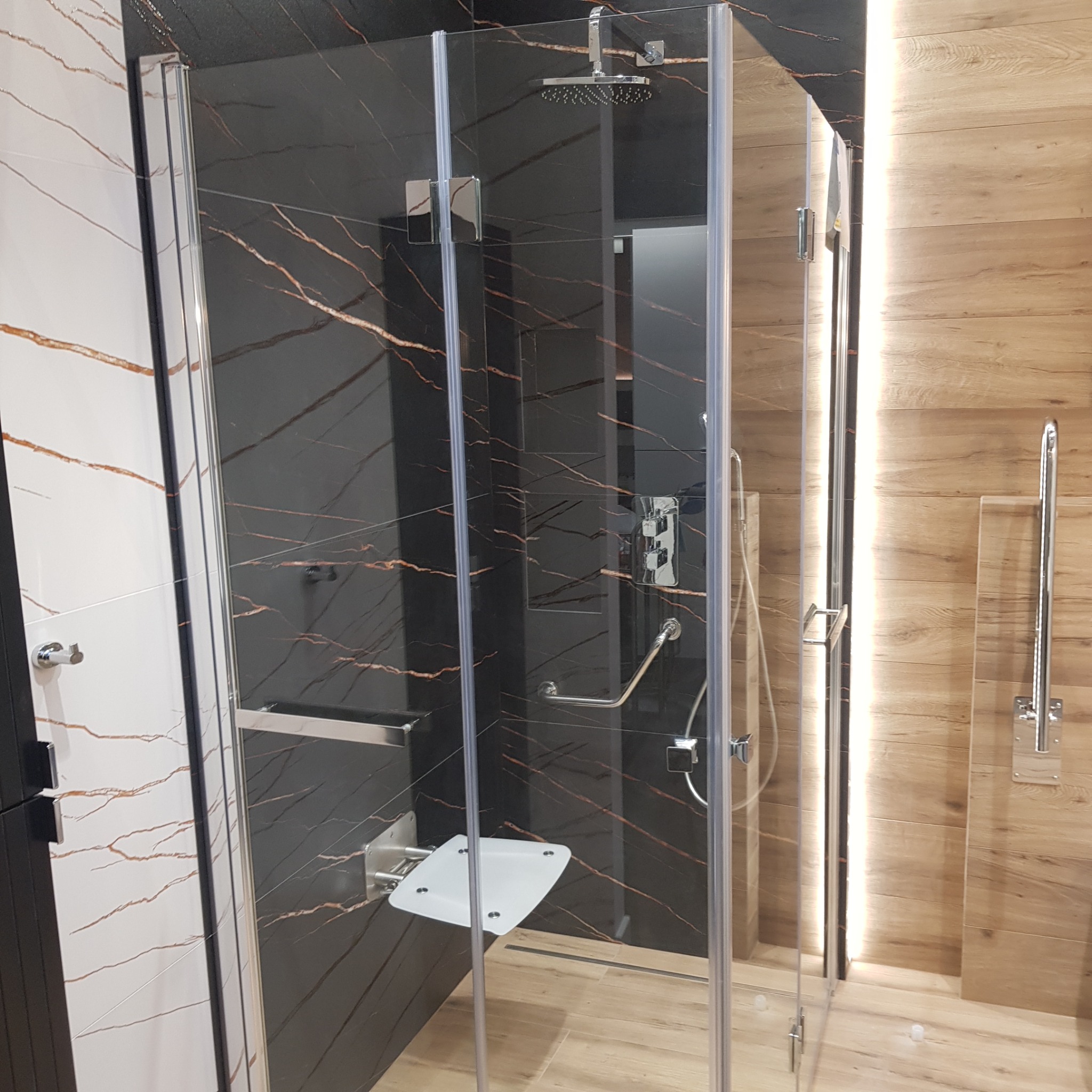 Nowoczesna kabina prysznicowa z siedziskiem i deszczownicą, wykończona płytkami imitującymi marmur i drewno, oświetlona subtelnym światłem.