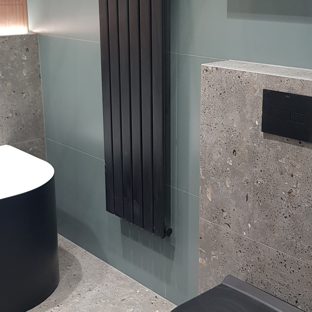 Nowoczesna łazienka: grafitowy grzejnik, ściany w odcieniach szarości i błękitu, minimalistyczny design. Umywalka częściowo widoczna, armatura w kolorze czarnym.