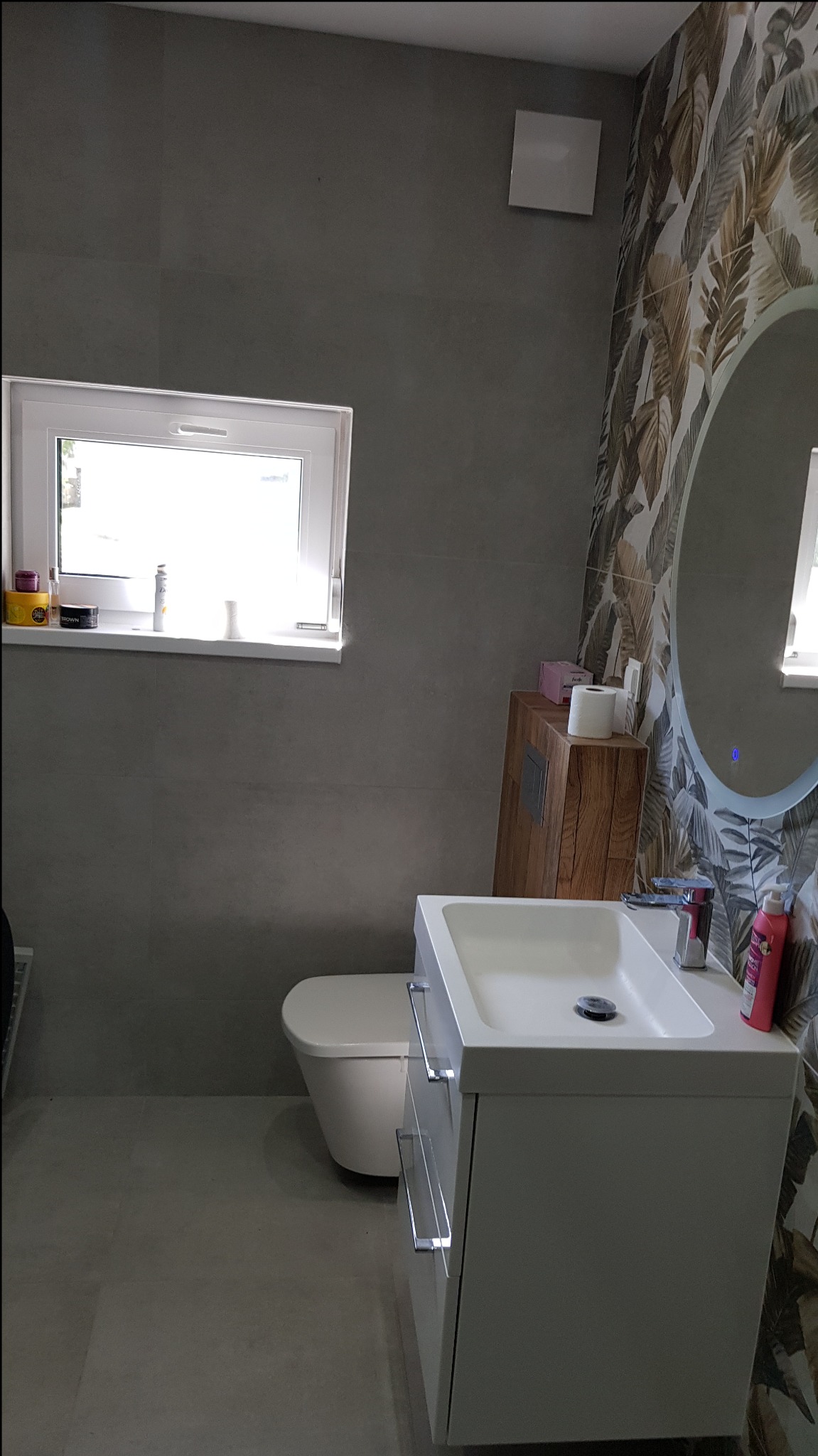 Nowoczesna łazienka z szarą płytką, wiszącą szafką z umywalką i lustrem z podświetleniem. Na ścianie wzorzysta tapeta z motywem liści.