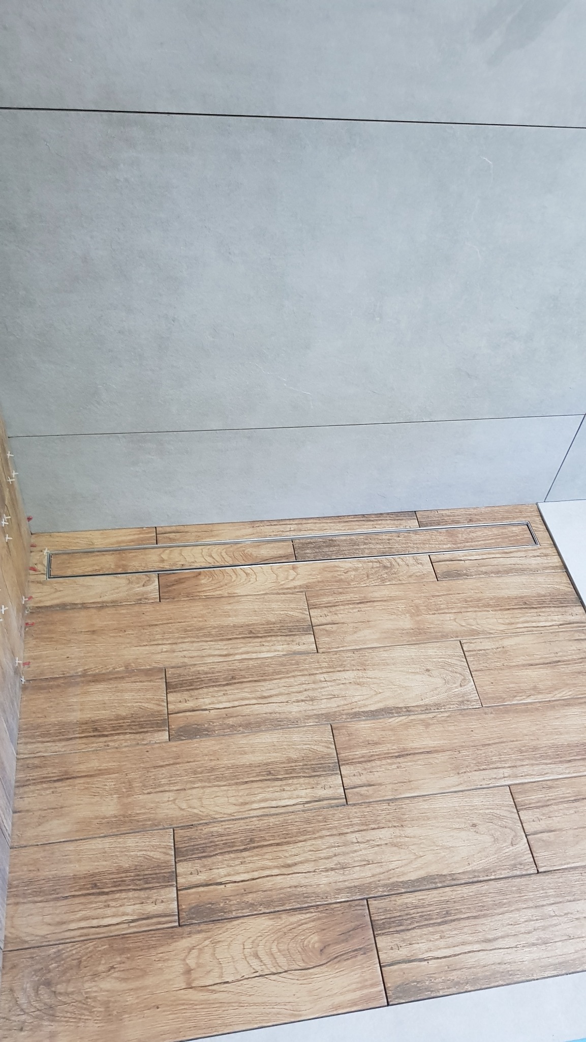 Ułożona podłoga z płytek imitujących drewno w łazience, z odpływem liniowym przy ścianie wyłożonej szarą, gładką płytką. Widoczne krzyżyki dystansowe.