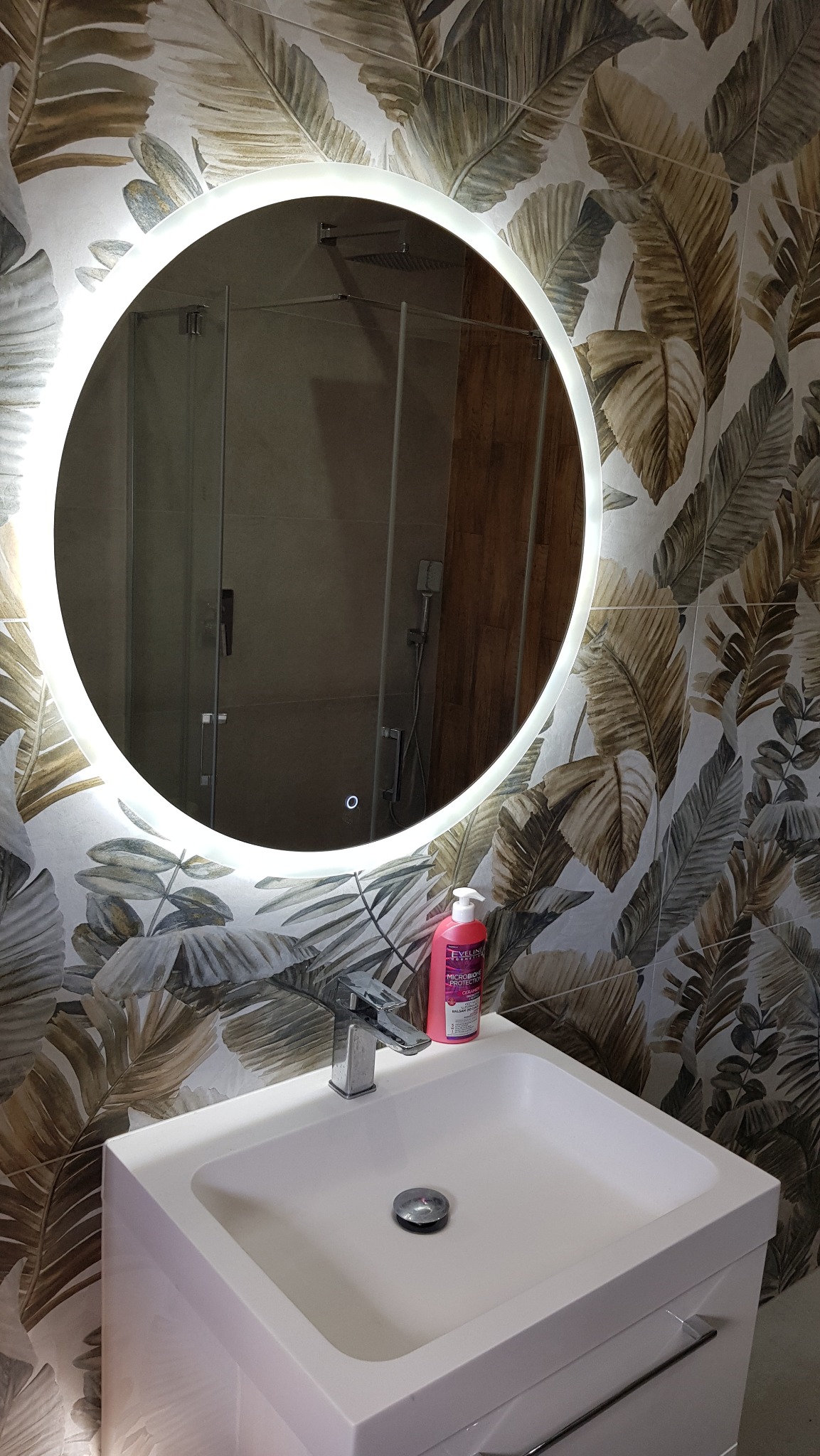 Nowoczesna łazienka z okrągłym lustrem LED i umywalką na tle ściany w liście bananowca. Widoczna kabina prysznicowa w odbiciu lustra.