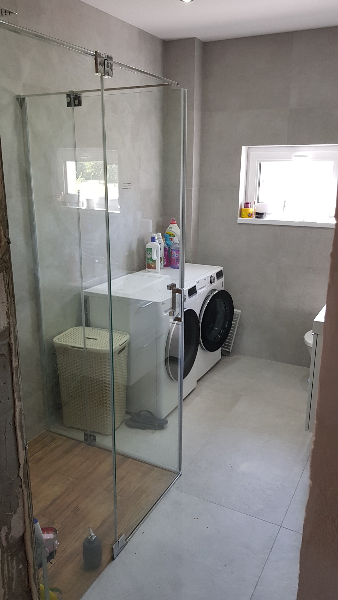 Wykończona łazienka z kabiną prysznicową ze szkła, pralką i koszem na pranie. Ściany i podłoga wyłożone płytkami w odcieniach szarości. Widoczne okno z dekoracjami.