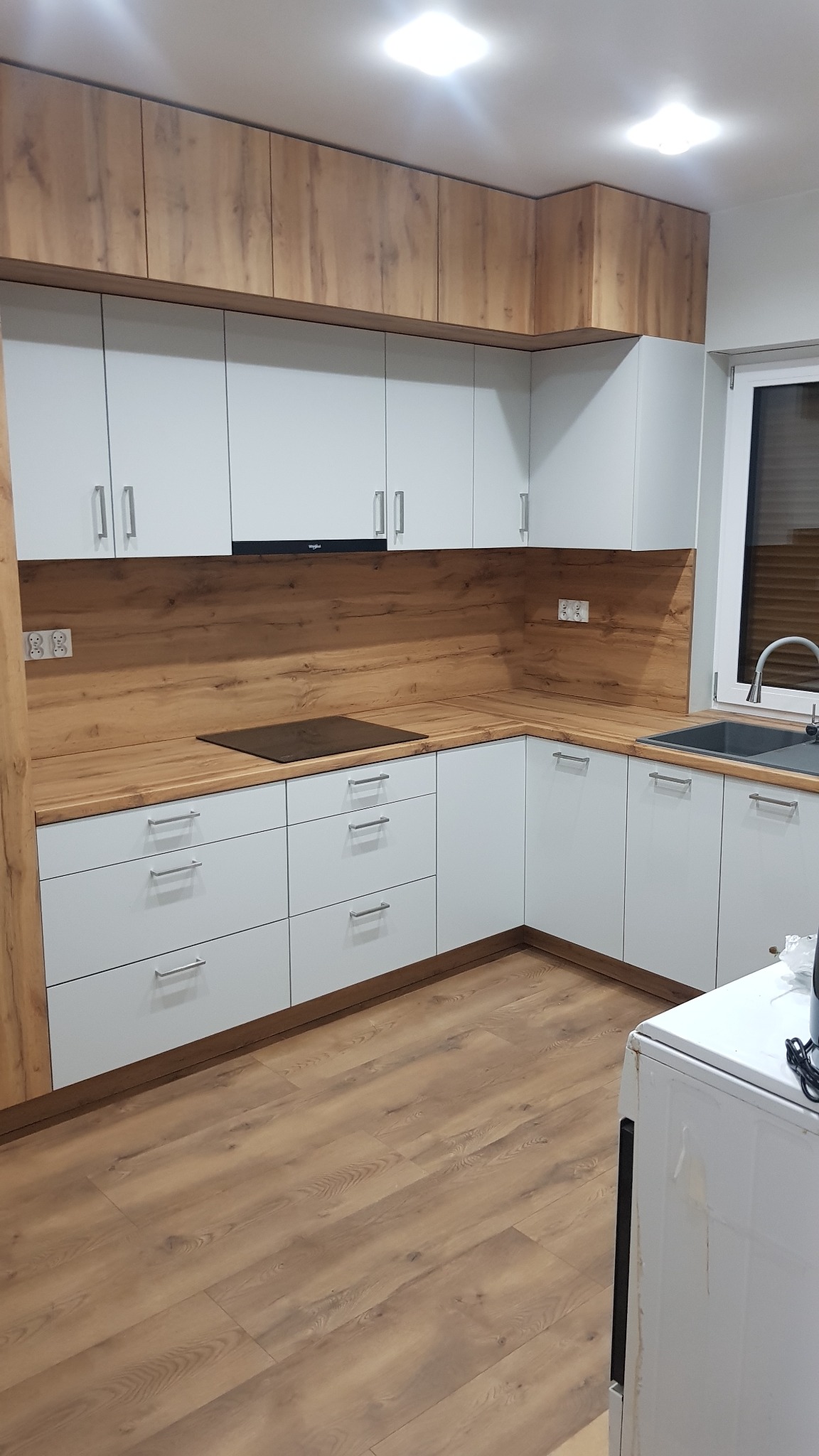 Nowoczesna kuchnia w odcieniach bieli i drewna z szarym zlewem, widok z perspektywy podłogi. Uwagę przyciągają drewniane szafki górne i blat.