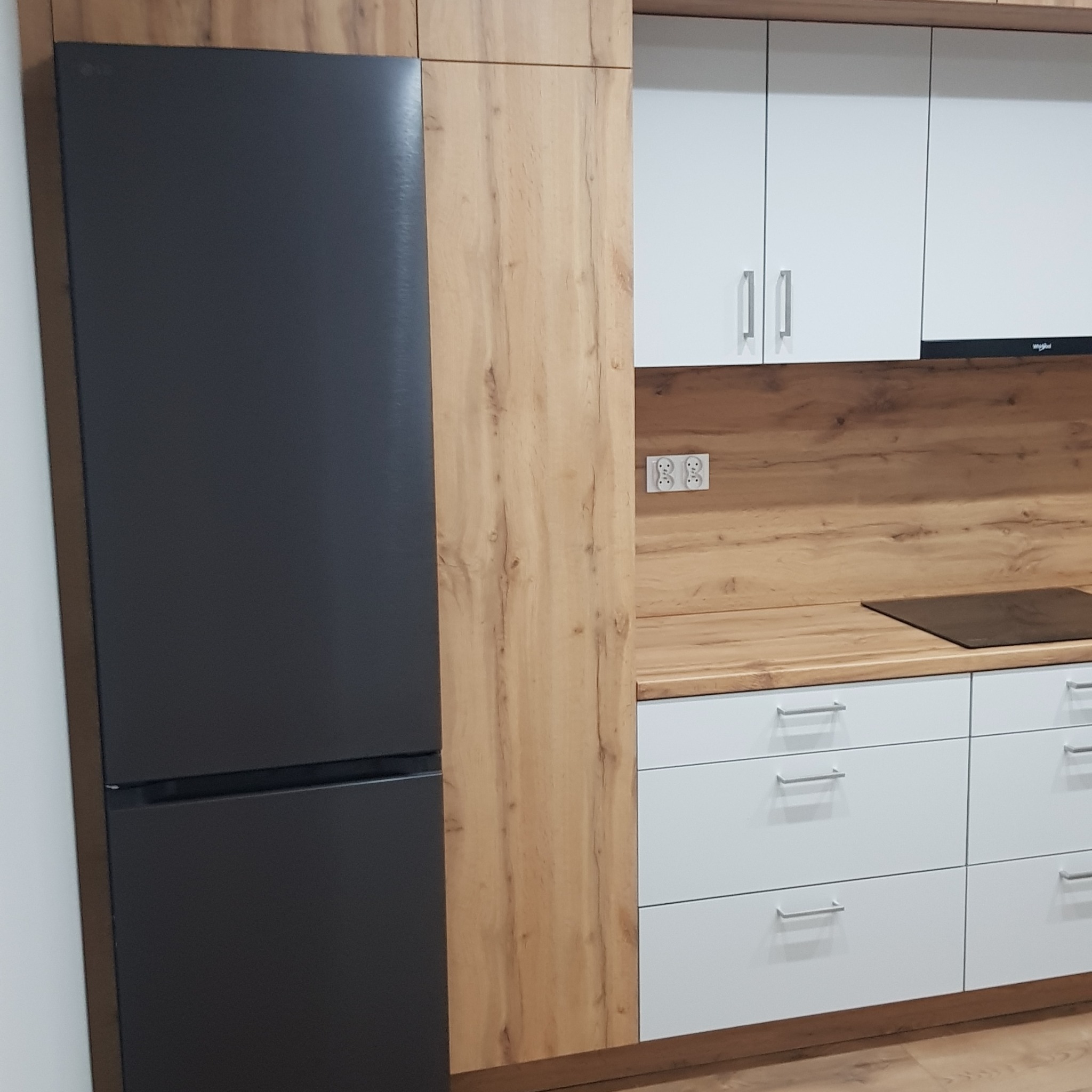 Nowoczesna kuchnia z szarym lodówką, drewnianymi panelami i białymi szafkami. Kontrastujące materiały tworzą elegancki wygląd. Widoczne gniazdka i płyta indukcyjna.