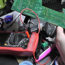 Libre Serwis - Lutowanie elementu na zielonej płycie drukowanej urządzenia elektronicznego, z użyciem czerwonego multimetru Unit-T, na zagraconym stole warsztatowym z widocznymi narzędziami i akcesoriami.