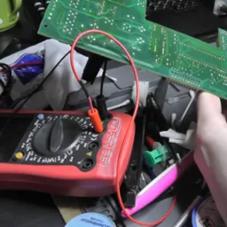 Lutowanie elementu na zielonej płycie drukowanej urządzenia elektronicznego, z użyciem czerwonego multimetru Unit-T, na zagraconym stole warsztatowym z widocznymi narzędziami i akcesoriami.